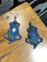 Pendientes de macramé azul oscuro con piedra central y cuentas doradas sobre mesa.