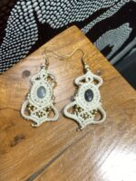 Pendientes de macramé blancos con piedra central ovalada y cuentas doradas, hechos a mano en India, sobre mesa.