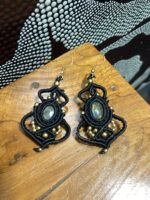 Pendientes de macramé negros con piedra central y cuentas doradas, artesanales, sobre mesa.