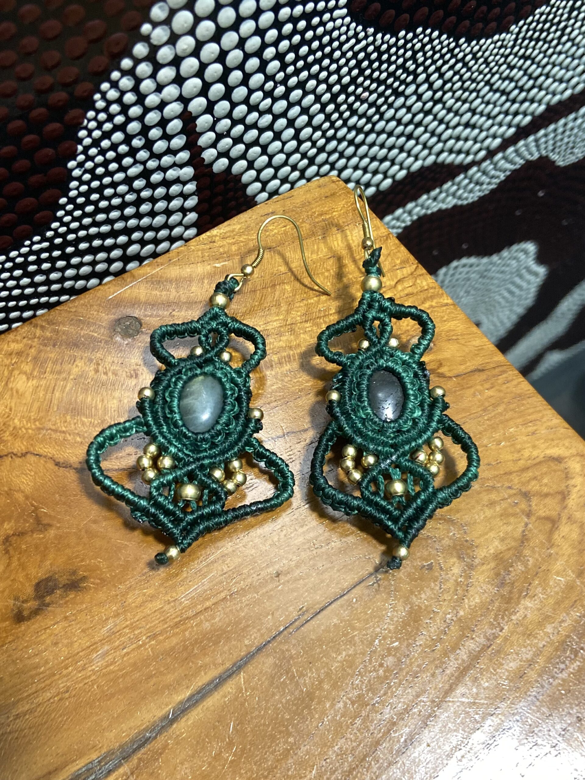 Pendientes en macramè verde con piedras Pendientes de macramé verdes con piedra central y cuentas doradas sobre mesa.